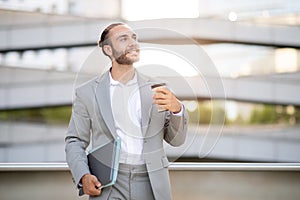 Smiling young businessman in suit holding laptop and takeaway coffee, walking outdoors