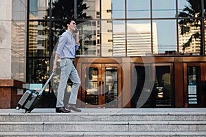 Smiling  Young Businessman Using Mobile Phone while Walking with Suitcase in the City. Lifestyle of Modern People