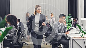 Smiling young businesswoman holding folder with papers and looking at coworkers while walking