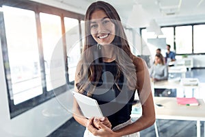 Smiling young businesswoman looking at camera while standing and holding her digital tablet in the modern startup office