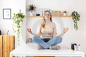 Smiling young businesswoman in lotus pose sitting on desk at home office