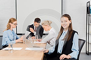 smiling young businesswoman sitting at modern office and looking at camera while colleagues having conversation