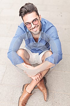 Smiling young casual man with glasses sitting on the sidewalk