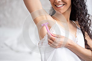 Smiling young caucasian brunette woman with long curly hair in pajamas shaves her armpit hair with razor