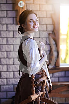 Smiling Young Caucasian Brunette Woman Posing With Fancywork Hoop in Front of Spinning Wheel in Retro Dress In Rural Environment