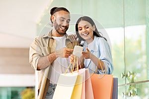 Smiling young caucasian couple with many packages enjoy walk outdoor, shopping, using smartphone app