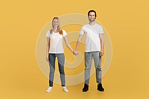 Smiling young caucasian female and male in white t-shirts hold hands, enjoy spare time, look at camera