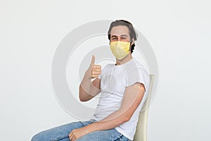 Smiling young caucasian man in protective mask with band-aid on shoulder after vaccination shows thumb up