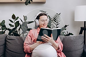 Smiling young Caucasian pregnant woman wear wireless headphone listen relax music and reading book to tell story to unborn child.