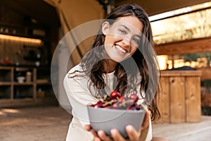 Smiling young caucasian woman holds out bowl cherries to camera spending leisure time outside city.