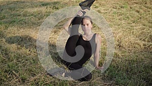 Smiling young Caucasian woman raising leg up and looking at camera. Portrait of flexible female gymnast sitting on grass