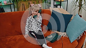 Smiling young children girl making conversation call on retro phone with friends on couch at home