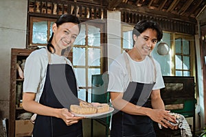 smiling young couple of cake makers wearing aprons holding up a tray of cooked cakes