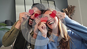 Smiling young couple covering their eyes with red heart shaped papers, having fun at Valentine Day. Boyfriend and