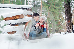 Smiling young couple with cups sitting in snow park. Drinking hot drinks, having fun.