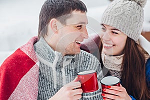 Smiling young couple with cups sitting in snow park. Drinking hot drinks, having fun.