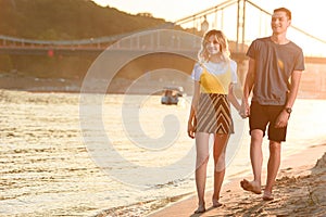 smiling young couple holding hands and walking barefoot on river beach