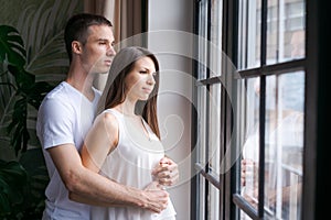 Smiling young couple hugging each other and standing near window, looking