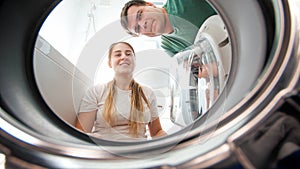 Smiling young couple looking inside washing machine and taking out clothes at laundry. Family doing housework and