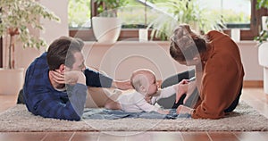 Smiling young couple lying together on rug on their living room floor at home with their adorable baby Baby looks at
