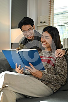 Smiling young couple relaxing on couch and reading book, spending leisure time on winter holidays at home