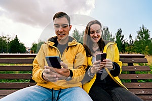 Smiling young couple in yellow jackets sitting on a bench in the autumn Park using phones, smartphones. Millennials, inseparable