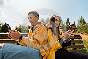 Smiling young couple in yellow jackets sitting on bench in autumn Park using phones, smartphones. Millennials, inseparable from