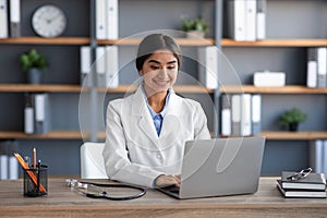 Smiling young cute indian woman medical worker, doctor in uniform typing on computer in office interior