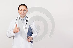 Smiling young doctor woman with stethoscope showing thumbs up isolated on white background. Female doctor in medical gown holding