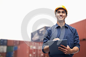 Smiling young engineer in protective work wear in a shipping yard examining cargo