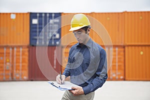 Smiling young engineer in protective work wear in a shipping yard examining cargo and writing on clipboard