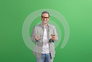 Smiling young entrepreneur paying bills online with credit card and mobile phone on green background