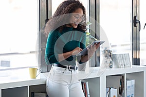 Smiling young entrepreneur woman using her digital tablet while standing next to the window in the office