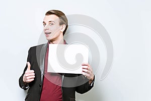 Smiling young european business man in red polo shirt and blazer holding blank A4 signboard and showing thumb up. Business