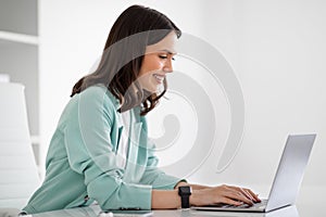 Smiling young european lady in suit at table typing on laptop, chatting with client