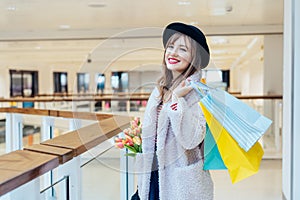 Smiling young fashion woman with net bag with fresh tulips flowers and paper shopping bags on mall background. Woman