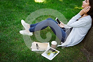 Smiling young female asian IT student in headphones. Listening to music and drinking coffee in the park