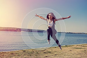 Smiling young female gymnast is jumping in split outdoors near the lake. Healthy lifestyle