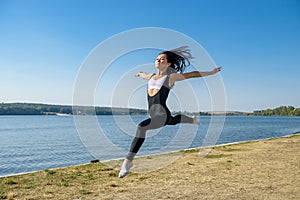 Smiling young female gymnast is jumping in split outdoors near the lake. Healthy lifestyle