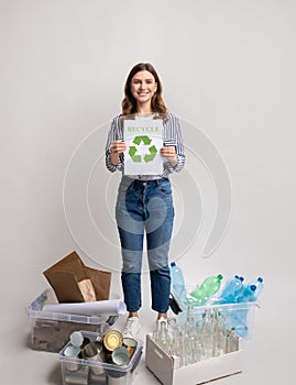 Smiling young female holding placard with recycle sign standing among sorted waste