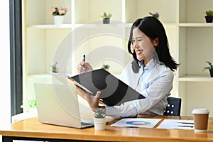 Smiling young female office worker sitting at desk and checking financial reports. Business, technology and