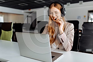 Smiling young female operator in headphones with headset working with laptop sitting at table at workplace, technical support