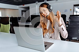 Smiling young female operator in headphones with headset working with laptop sitting at table at workplace, technical support