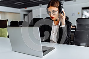 Smiling young female operator in headphones with headset working with laptop sitting at table at workplace, technical support