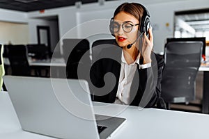 Smiling young female operator in headphones with headset working with laptop sitting at table at workplace, technical support