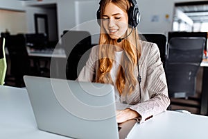 Smiling young female operator in headset works while sitting at the table, uses laptop, working in the call center