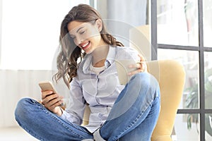 Smiling young female sitting in the armchair in the living room and using with her mobile phone