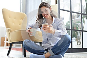 Smiling young female sitting on the floor in the living room and using with her mobile phone