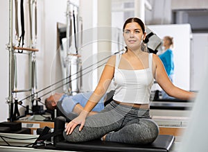 Smiling young female in sportswear training twist rotation while using Pilates performer bed in gym