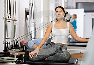 Smiling young female in sportswear training twist rotation while using Pilates performer bed in gym